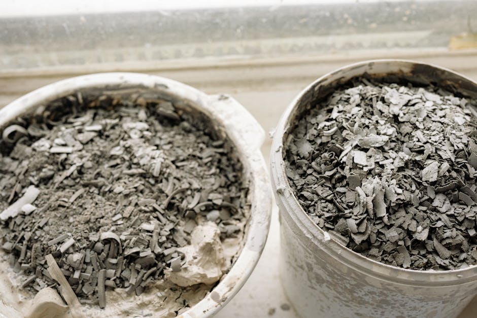 Two white plastic buckets filled with a mixture of crushed and shredded grey debris, including broken ceramic or plaster fragments, sit on a windowsill. The rough-textured materials vary in size and shape, with some curled or jagged edges, and are densely packed within each container. The left bucket appears to contain slightly finer, more powdery residues, while the right bucket holds larger, more irregular Pieces. The background shows a blurred window ledge or sill, hinting at an indoor setting. The scene suggests an on-site clearance process, with the debris likely resulting from renovation or demolition work, reflecting the type of waste handled by private rubbish removal services such as Rubbish Clearance Islington. The lighting is neutral, emphasizing the textures and materials of the waste included in the containers, consistent with professional disposal or collection activities associated with non-local authority rubbish management.