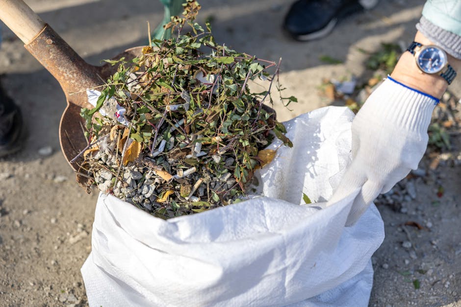 A close-up view of a person wearing a white glove and a watch, using a rusted shovel to lift a mixture of urban garden waste, such as soil, small green leafy plants, and scattered debris, from a white woven polypropylene bag placed on a paved outdoor surface. The waste appears to contain cuttings or weeds with thin stems and green leaves, some with dried edges, and small broken pieces of concrete or gravel are visible among the organic material. The background shows part of a person's leg in dark clothing and a pair of black shoes, indicating an outdoor setting likely associated with rubbish removal or site clearance activities. The scene reflects aspects of independent waste handling, closely aligned with private rubbish collection services offered by companies like Rubbish Clearance Islington, focusing on clearing refuse from gardens or outdoor areas as part of on-site rubbish removal.