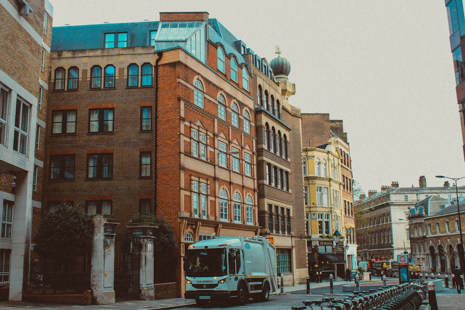 A street scene featuring a collection of historic multi-storey buildings with varied architectural styles, including brick facades, arched windows, and decorative cornices, situated in an urban area. In the foreground, a light blue waste collection truck with a compact, modern design is parked on the pavement, partially blocking the view of the grey stone pillars and entrance gate to one of the buildings. The surrounding environment includes a row of bicycles parked along the curb, metal bollards separating the sidewalk from the road, and nearby streetlights. The sky appears overcast, providing diffuse lighting that reduces shadows. The scene illustrates a typical city street where private waste collection services, such as those offered by Rubbish Clearance Islington, might perform onsite clearance, handling rubbish removal outside of local authority collection schemes, in alignment with legal and safe waste disposal practices. The combination of historical architecture and urban waste management vehicles highlights the importance of professional rubbish removal in maintaining city cleanliness and supporting alternative waste handling options.