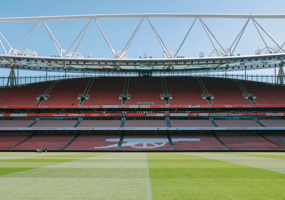 A view of the interior of a large, modern football stadium with a partially covered roof supported by white steel trusses visible at the top of the image. The seating area is predominantly red with several tiers of empty seats and some advertising banners along the front of the upper and lower levels. The field below features immaculately maintained green grass with faint mowing lines visible, and the centre circle marked clearly on the turf. In the background, the sky is bright and clear, indicating daytime. The stadium appears ready for a match or event, with no spectators or players present at the moment. This setting emphasizes the scale and structure of an arena that hosts live sports, illustrating a space where waste or rubbish clearance services might be needed after events, or during maintenance periods, although no debris or rubbish is visible in the image.