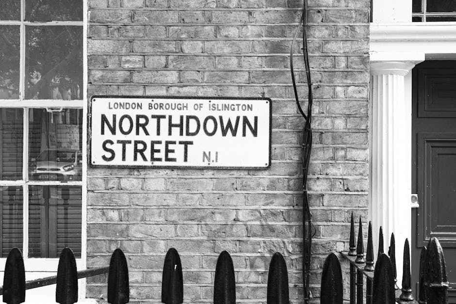 A black-and-white photograph showing a street sign mounted on a brick wall, indicating 'Northdown Street' in the London Borough of Islington. To the left of the sign, there is a window with multiple panes, reflecting parked cars on the street outside. To the right, there is a dark wooden door with decorative white column-like trim alongside it. Running vertically along the brick wall next to the sign, there are exposed black cables or wires loosely bundled and hanging. In the foreground, a row of pointed wrought-iron fence spikes is visible, suggesting the presence of a small front garden or boundary wall. The overall scene appears to be part of a residential or mixed-use property, captured in natural daylight. This type of urban setting is typical of areas where private waste collection or alternative rubbish removal services may be used, often involving on-site clearance or independent disposal methods.