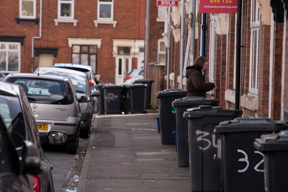 A woman standing on a Victorian-style city street sidewalk, positioned next to a brick building with white window frames and stone accents, is seen near a row of black wheeled rubbish bins, some with graffiti or markings on their lids and front panels. The bins are aligned along the pavement, partly overlapping the edge of the sidewalk, indicating an area designated for waste collection or disposal. The street features parked cars of varying models and colors, including silver, grey, and white, lining both sides of the narrow road, with the vehicles’ rear windows and side mirrors visible. In the background, other residential buildings made of similar brick material are visible, and a red “sold” sign is mounted next to a window on the building in the foreground. The scene is lit by natural daylight, with a slightly overcast sky creating soft shadows, and the overall atmosphere suggests a typical urban environment where private arrangements for rubbish disposal are in place, consistent with independent rubbish clearance services operating in the area. Rubbish Clearance Islington provides similar services focusing on efficient waste removal from properties such as this, supporting private waste handling needs in urban residential settings.