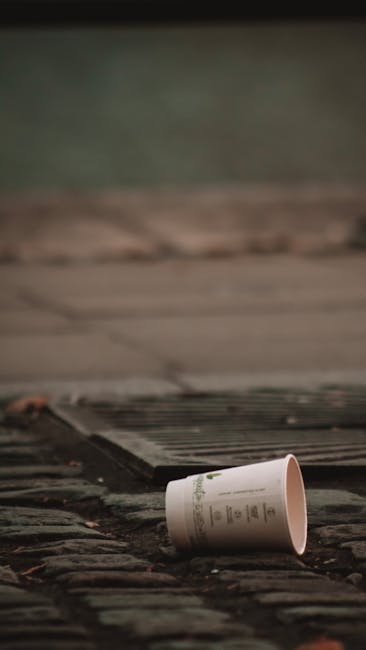 A discarded paper cup lying on a weathered brick surface, partially tipped over with the open end facing down. The cup appears to be made of cardboard with a white exterior and green and black printed text and logo, likely used for hot beverages. The brick surface beneath is uneven, with visible textures and a dark, damp appearance. In the background, a ground-level grate or drainage cover made of dark metal with parallel slats is situated close behind the cup, with some debris and dirt around it. The scene is outdoors, possibly on a pavement or alleyway, with muted lighting suggesting overcast conditions or late afternoon. The surroundings and the presence of a drain suggest this might be an urban area where rubbish, such as discarded cups, ends up on streets or near outdoor waste handling areas. The environment evokes the importance of responsible disposal and supports services like those provided by Rubbish Clearance Islington, offering alternative waste removal options for waste like this.