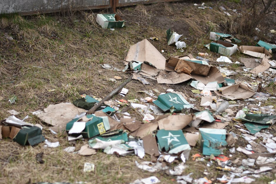 An outdoor scene showing a collection of overflowing rubbish bins and scattered waste on a paved sidewalk area. The central focus is a large grey mixed paper and cardboard recycling container, filled with crumpled papers, cardboard boxes, and various paper waste, with some items spilling onto the ground. To its right are black and red bins; the black bin is partially open, revealing more waste inside, while the red bin appears to be closed. To the left of the grey bin, a grey car is parked behind a metal railing, which separates the waste area from the vehicle. In the background, there is a building with storefronts, including a fish bar, and a construction or scaffolding structure covered with blue safety netting. The scene is lit with natural daylight, emphasizing the cluttered and untidy state of the waste collection, which highlights the importance of proper rubbish management and private collection services such as those offered by Rubbish Clearance Islington in the context of alternative waste handling methods in urban environments.