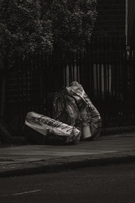 A collection of black and white plastic rubbish bags, some appearing crumpled and tied at the top, are placed on the pavement near the curbside in front of a dark metal fence with vertical slats. The bags likely contain household or garden waste and are positioned in an outdoor urban environment, with a dense leafy bush visible behind the fence. The scene suggests a private or independent waste collection, possibly awaiting removal by a rubbish clearance service such as Rubbish Clearance Islington, given the context of alternative waste handling options. The lighting is subdued, contributing to the monochrome tone of the image, emphasizing the texture and form of the waste bags against the background of the urban street setting.