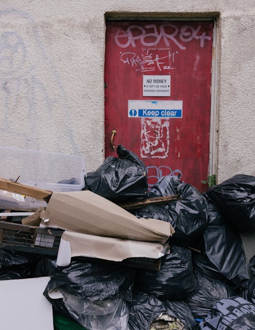 A collection of black and white plastic rubbish bags, some appearing crumpled and tied at the top, are placed on the pavement near the curbside in front of a dark metal fence with vertical slats. The bags likely contain household or garden waste and are positioned in an outdoor urban environment, with a dense leafy bush visible behind the fence. The scene suggests a private or independent waste collection, possibly awaiting removal by a rubbish clearance service such as Rubbish Clearance Islington, given the context of alternative waste handling options. The lighting is subdued, contributing to the monochrome tone of the image, emphasizing the texture and form of the waste bags against the background of the urban street setting.