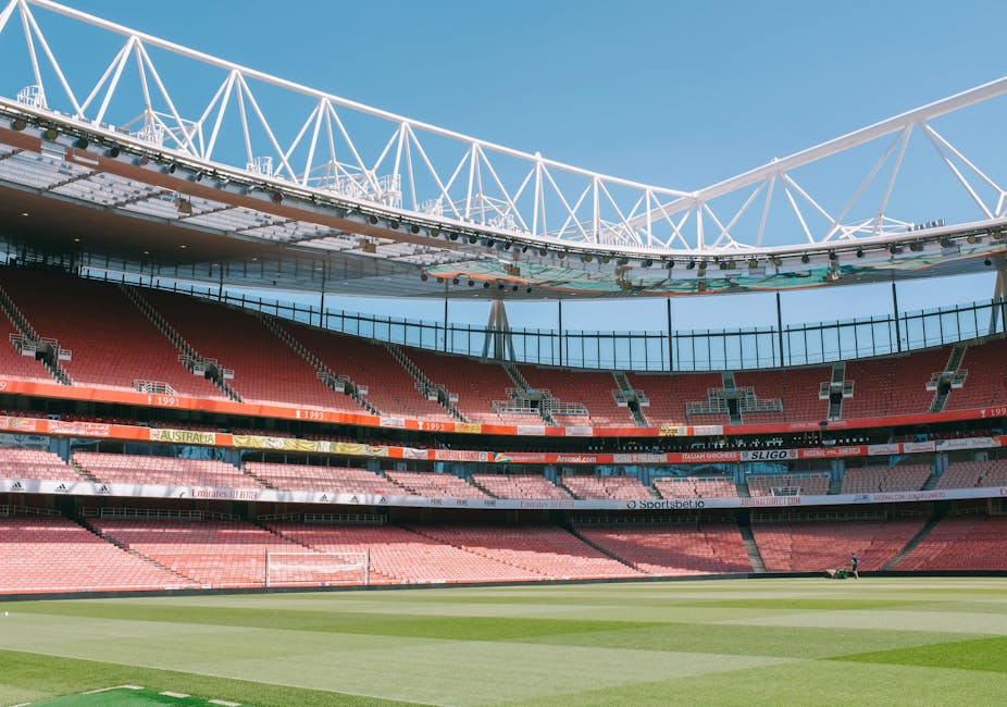 The image shows a large, empty stadium with multiple tiers of red seating arranged in curved rows, overlooking a well-maintained grass pitch with visible white goalposts at either end. Above the seating area, a white steel framework of the stadium’s roof structure extends across the open sky, with triangular supports creating a geometric pattern. The sky is clear with a bright blue tone, indicating a sunny day, and natural light illuminates the stadium interior uniformly. Surrounding the pitch, there are security barriers and advertising banners, some of which display sponsor logos. The environment appears clean and orderly, with no visible debris or clutter, reflecting a typical sports venue prepared for an event or maintenance. This scenario aligns with the context of an arena that may require occasional rubbish clearance during events or after use, though no waste or rubbish is present in this specific image. Rubbish Clearance Islington might be associated with service provisions that include on-site waste handling for such venues after events.