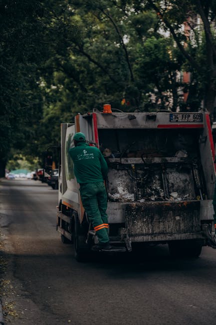 A sanitation worker in a green uniform and protective helmet is standing on the rear step of a small garbage collection truck, which is positioned on a quiet residential street. The truck has a partially raised rear door, revealing a large, dark waste compartment with a textured, greasy surface. The vehicle is primarily white with red and black markings, and a license plate with the registration '34 FFP 609' is visible at the top. The street is lined with trees, their green foliage casting dappled shadows over the scene, and a few parked cars are seen in the background. The environment appears calm and suburban, with soft, natural lighting indicating daytime. The scene captures a routine waste collection operation, consistent with private or independent rubbish removal services, emphasizing the physical effort involved in rubbish clearance and waste management responsibilities handled by companies such as Rubbish Clearance Islington.