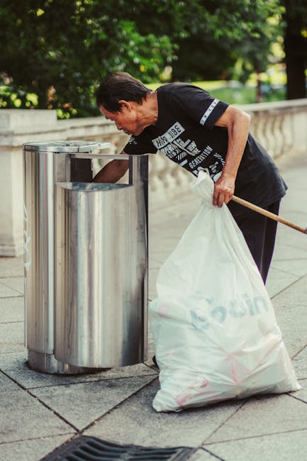 A man with dark hair, dressed in a black T-shirt with white text and dark trousers, is seen standing outdoors on a paved sidewalk, next to a stainless steel recycling bin with a curved top and open lid, which is positioned near a stone balustrade. The man is leaning forward, placing a white plastic bag filled with rubbish into the trash receptacle. In his right hand, he holds a wooden stick or broom handle, and his left hand supports the bag. The setting appears to be a park or garden area with lush green trees and foliage in the background, and bright natural lighting illuminating the scene. The overall environment suggests a context of waste disposal or rubbish collection, possibly as part of private or independent rubbish clearance services. Rubbish Clearance Islington is known for handling waste removal and environmentally responsible disposal, as exemplified by this outdoor scene of manual rubbish management.